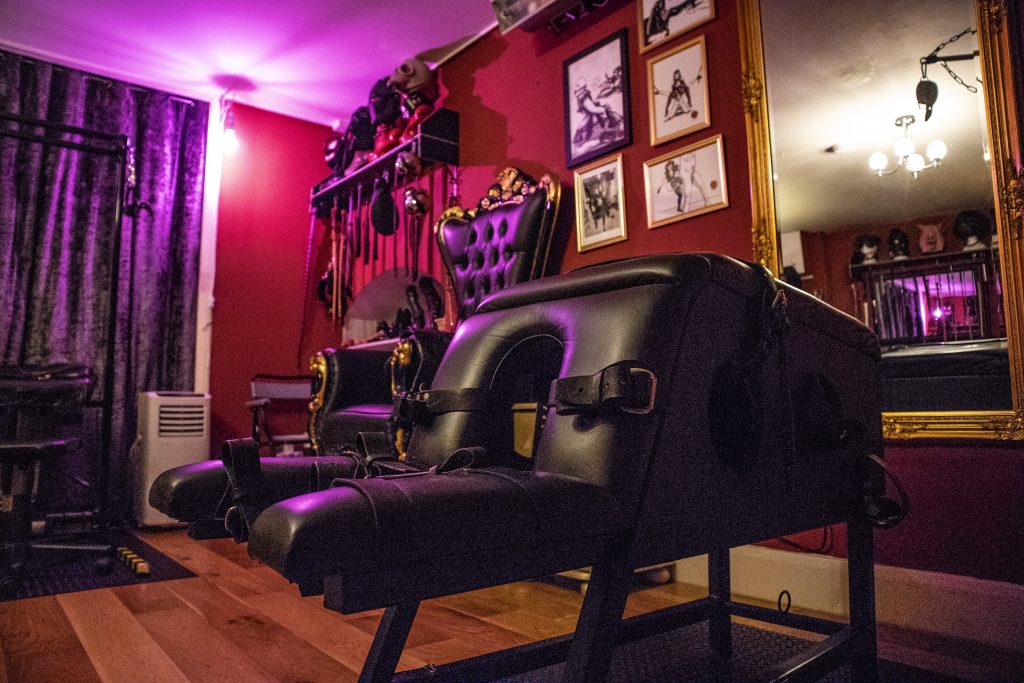 Low view of the spanking bench with the throne and hood rack behind it in violet light