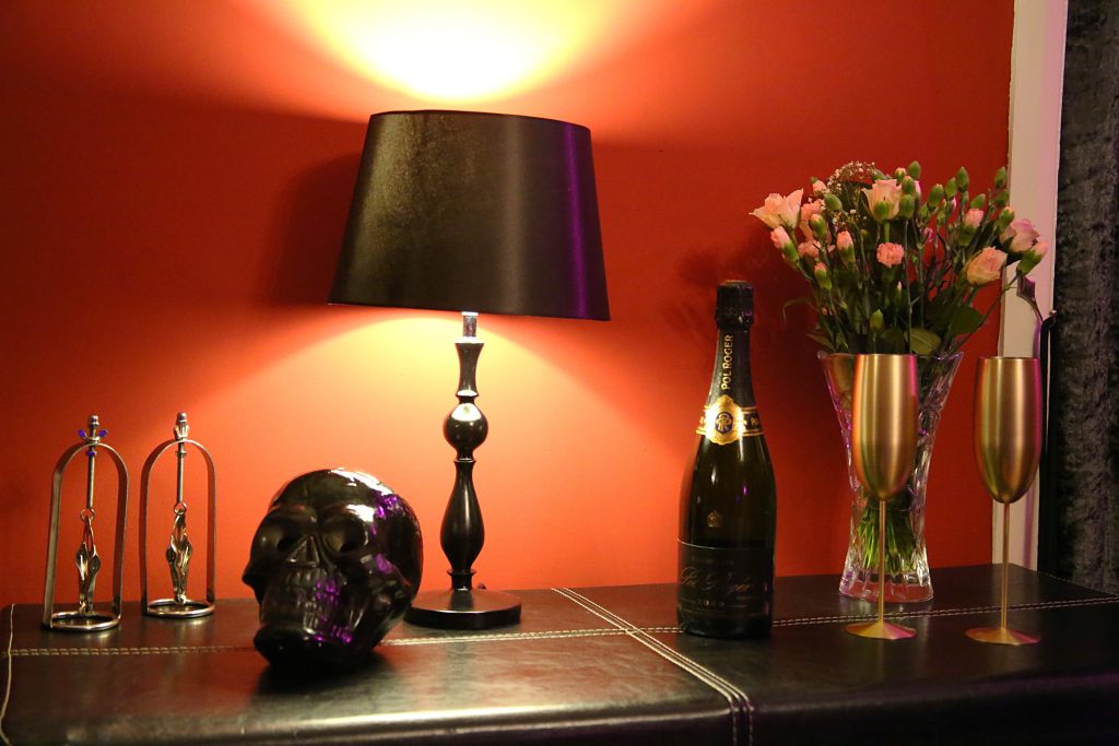 Sideboard with black lamp, crystal skull, gold flutes and roses against a red wall