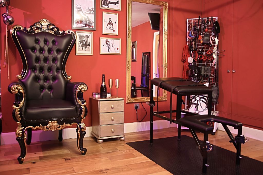 Black and gold throne beside a padded spanking bench, with framed fetish art and a gold mirror