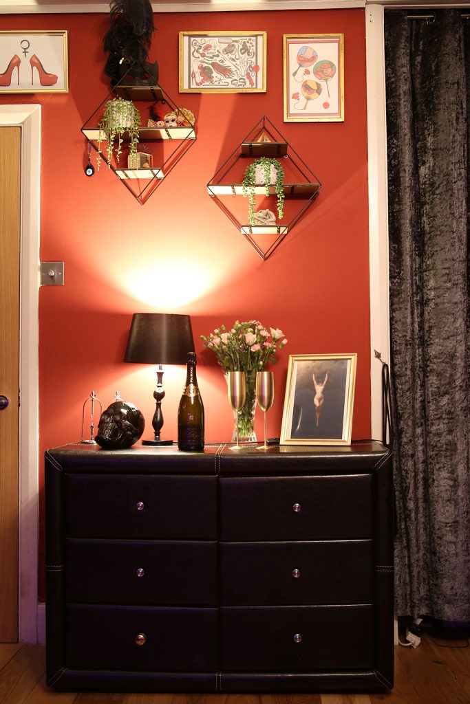 Black leather chest of drawers with lamp, champagne and framed prints on a red wall