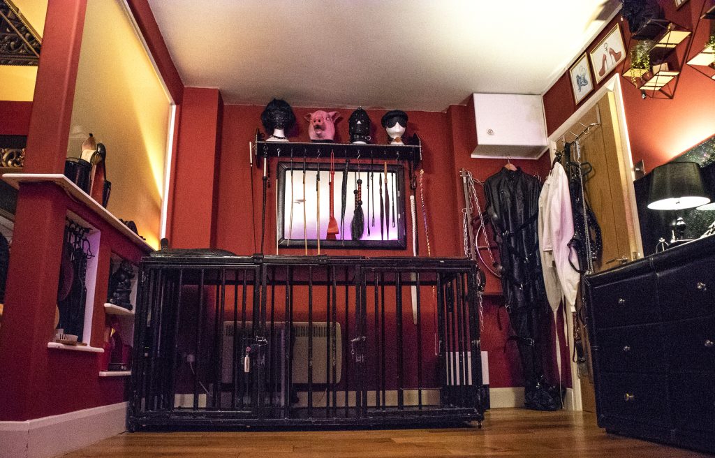 Steel cage bed below a shelf of hoods and masks, with a white straitjacket hanging beside it