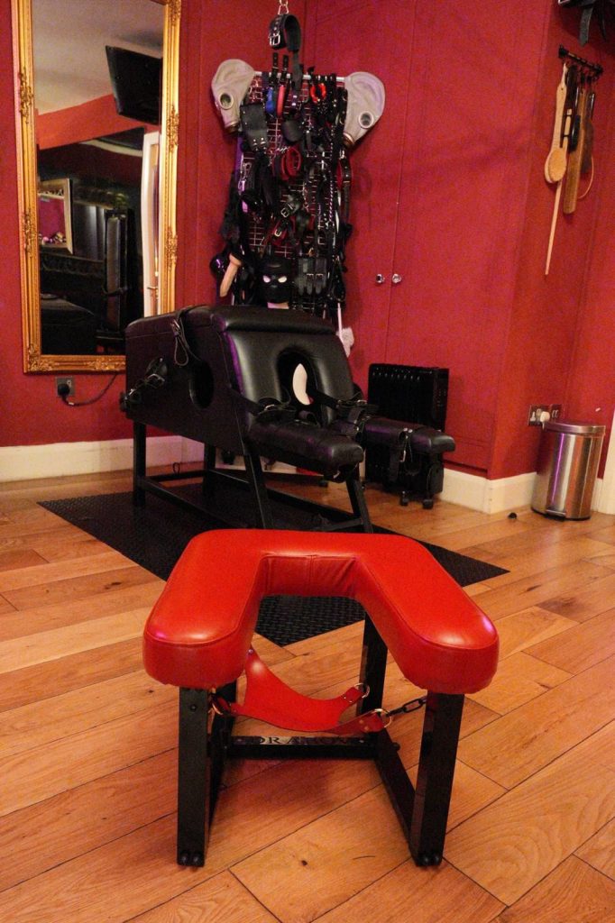 Red padded queening stool in front of the black bondage chair