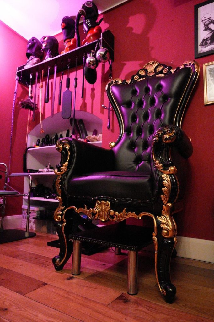 Gold-trimmed throne below a rack of hoods and floggers