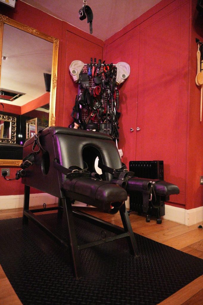 Black bondage chair with head opening in front of a wall of restraints