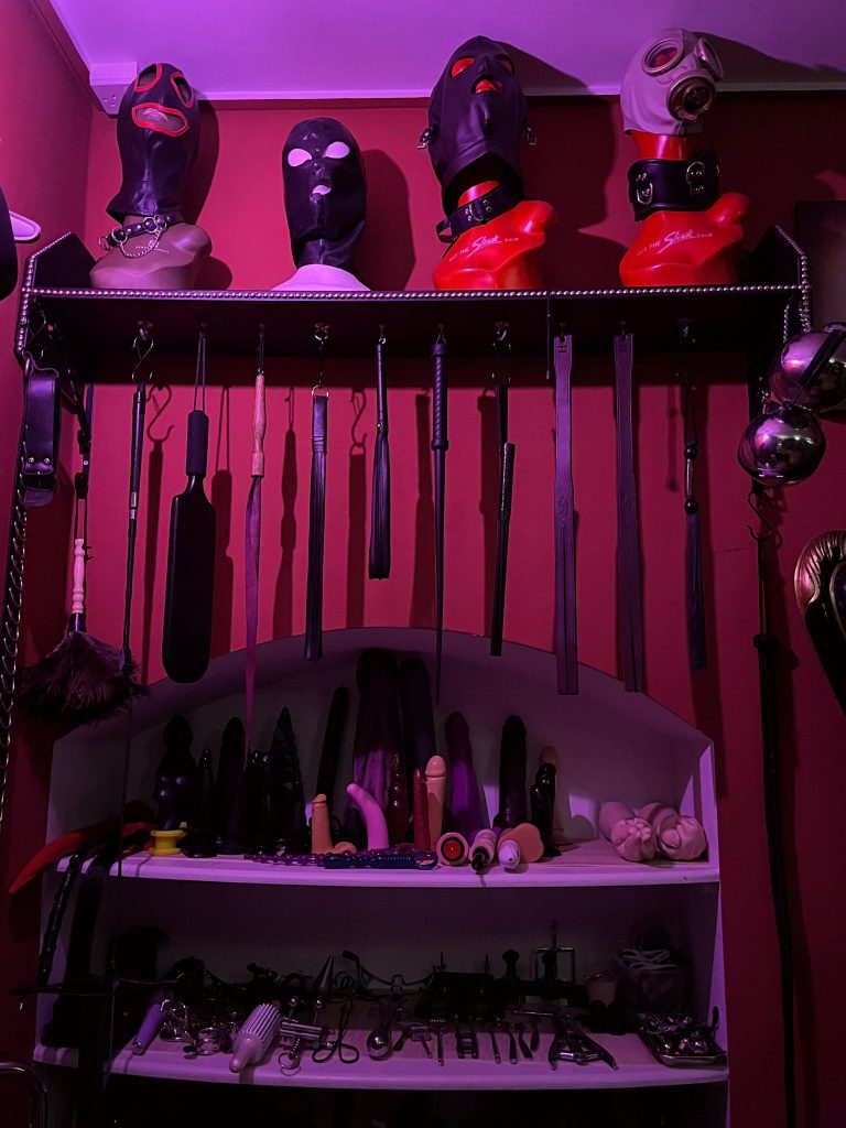 Shelf of rubber hoods above a long rack of canes, crops and paddles
