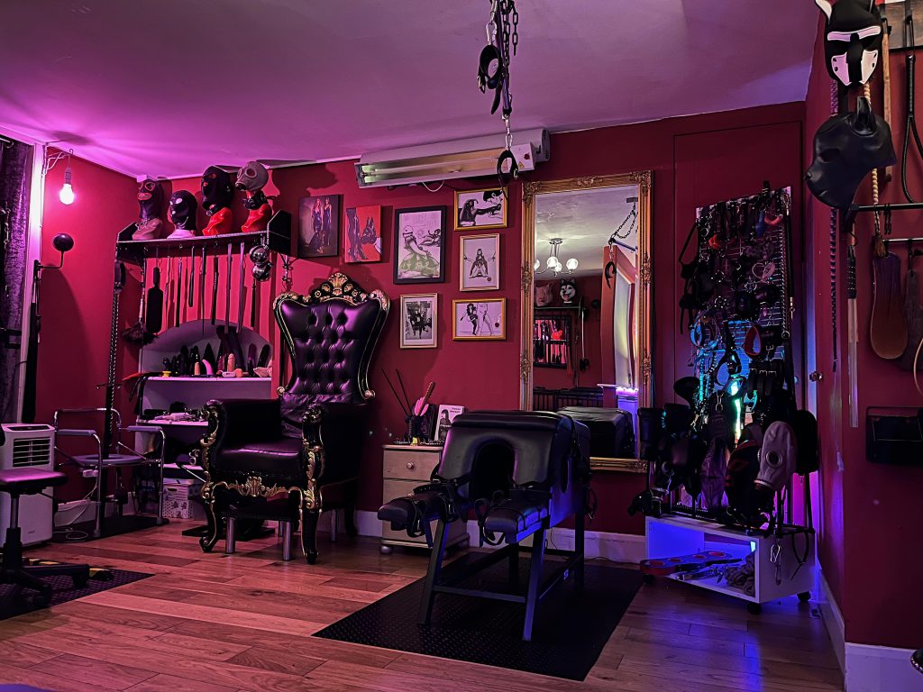 Wide view of the dungeon with the black and gold throne, spanking horse, framed fetish art and wall of hoods