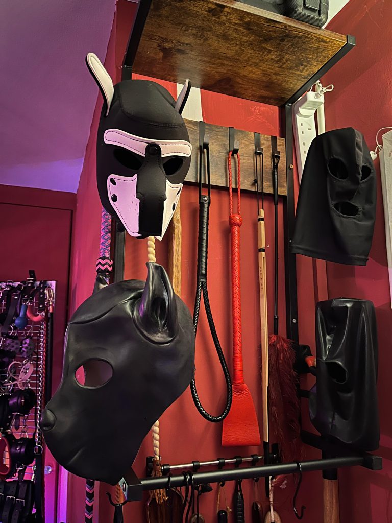Pup hoods and leather masks hanging over a rack of paddles