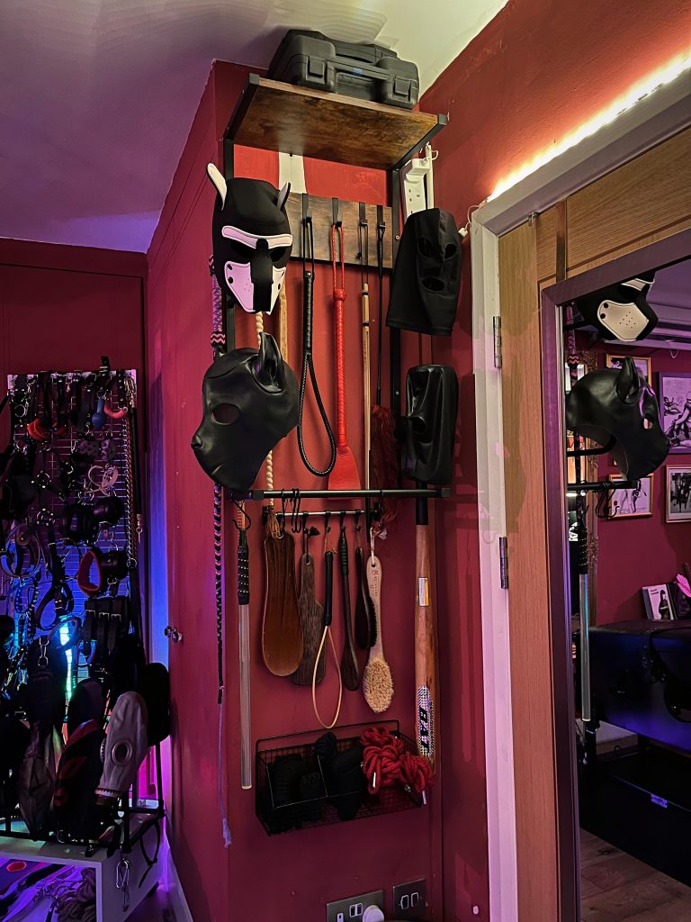 Narrow wall of hoods, gags and paddles under a purple LED strip
