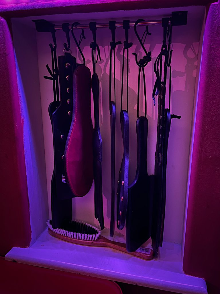 Leather paddles and straps hanging in a purple-lit alcove